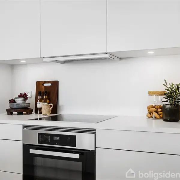 Induktionskogeplade i moderne hvidt køkken omgivet af skabslåger. På bordet er en skærebræt med flasker og skåle med frugt, samt en glasbeholder med småkager og en plante.