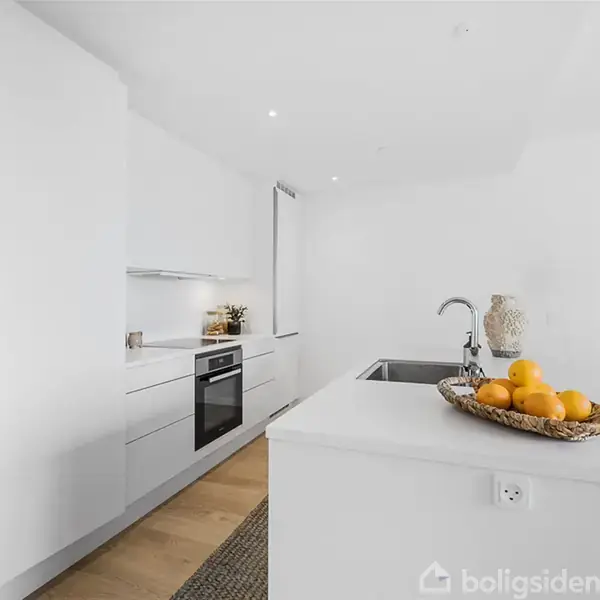 Køkkenbord med frugtskål fyldt med appelsiner står centralt; moderne hvidt køkkenmiljø med indbygningsovn, underskabe, og vask. Trægulv skaber varme i det minimalistiske rum.