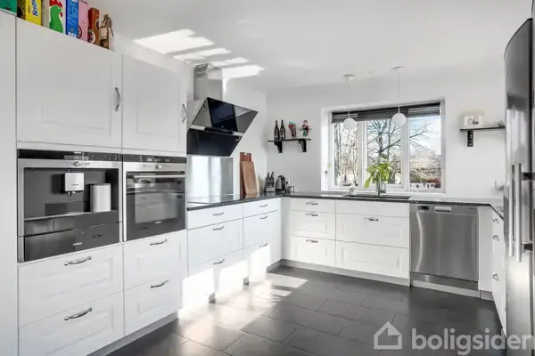 Et moderne køkken med hvide skabe og skuffer, der indeholder indbyggede apparater som ovn og kaffemaskine. Rummet har et stort vindue med udsigt til træer udenfor.