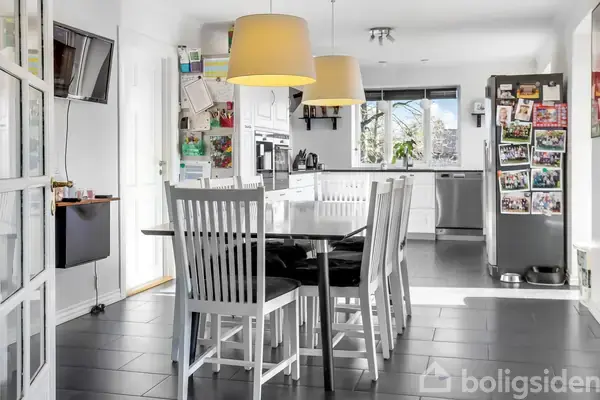 Et spisebord med hvide træstole står i forgrunden. To lamper hænger over bordet. Baggrunden viser et moderne køkken med hvide skabe, sort bordplade og et køleskab fyldt med magneter.