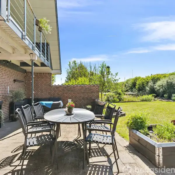 Rundt havebord med seks stole står på en terrasse foran et murstenshus. I baggrunden er en grøn have med buske og et bålsted under en blå himmel.