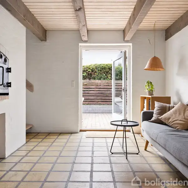 En moderne stue har en grå sofa med puder, et lille sofabord og en væghængt pejs med brænde under. En glasdør fører ud til en terrasse med grønne buske.