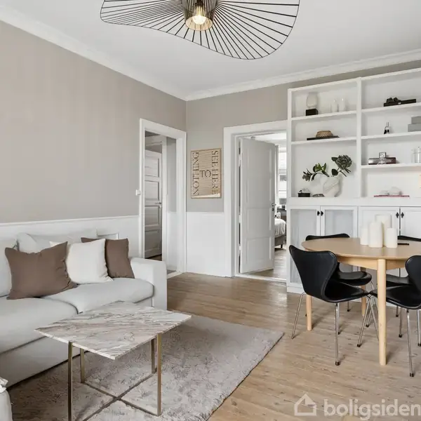 En lys stue med en grå sofa placeret ved siden af et marmorbord. En rund træspisebord med fire sorte stole er i nærheden. Indbyggede hylder og en åben dør i baggrunden.