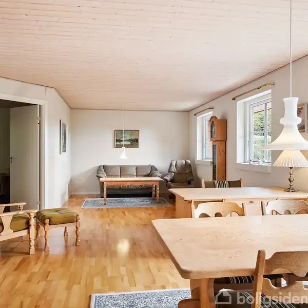 En lys stue med trægulv har et spisebord med stole i forgrunden, to sofaer i baggrunden, og kunst på væggene, omgivet af store vinduer med udsigt til haven.