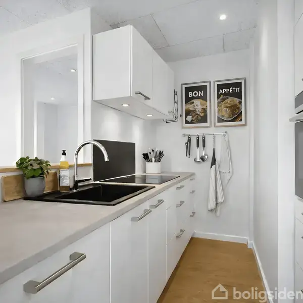 Et lyst køkken med hvide skabe og en sort vask. Organiserede redskaber hænger fra væggen. Over vasken er to plakater med teksten "BON" og "APPÉTIT".