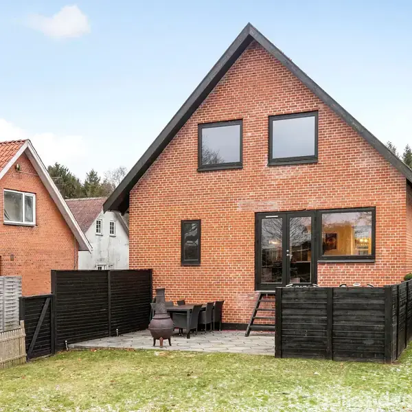 Rødstenshus med sort tag står stille i en have med terrasse, indhegnet af sorte hegn. Til venstre ses et andet hus, og en båd er synlig til højre.