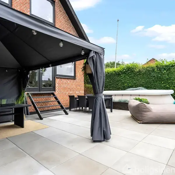 Pavillon med gardiner står på flisebelagt terrasse. Ved siden af er en stor pude og en oppustelig pool. Omgivet af hæk, ses en muret bygning i baggrunden.
