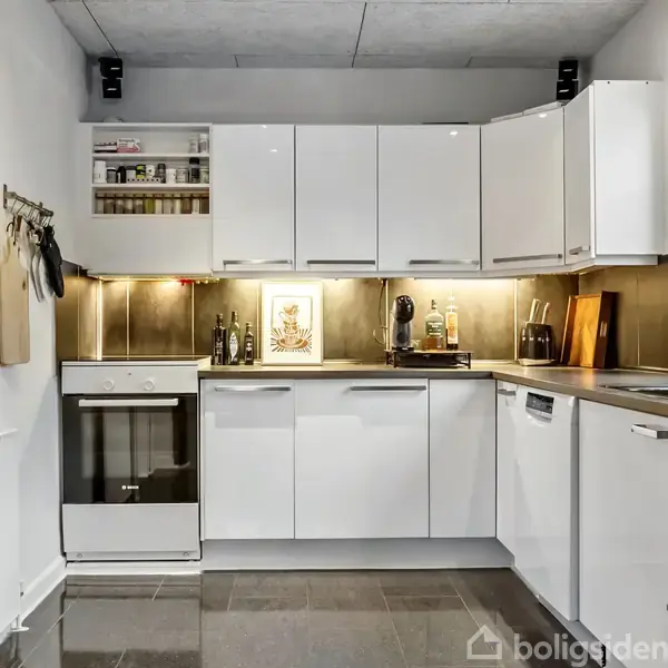 Hvidt køkken med moderne skabe, opvaskemaskine, og ovn indbygget. Pyntegenstande og køkkenredskaber er placeret på bordplade og hylder. Væggene har dekorative elementer.