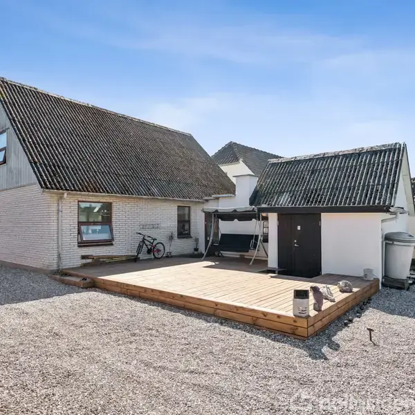 Hus med skråtag står på en grusbelagt grund. En træterrasse foran med cykel og bænkesæt. Gård omgivet af hække og naboens tag ses i baggrunden.