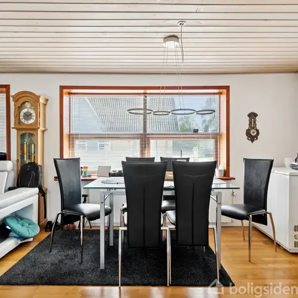 Et spisebord med seks sorte stole står på et sort tæppe i en lys stue. En hvid sofa står til venstre ved siden af et ur. Vinduerne har persienner.