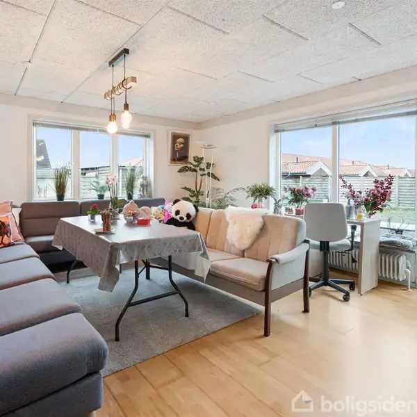 Grå sofa og bord med dug fyldt med pyntegenstande i en lys stue. Store vinduer giver udsigt til haven med græsplæne. Venstre væg har billeder og planter.