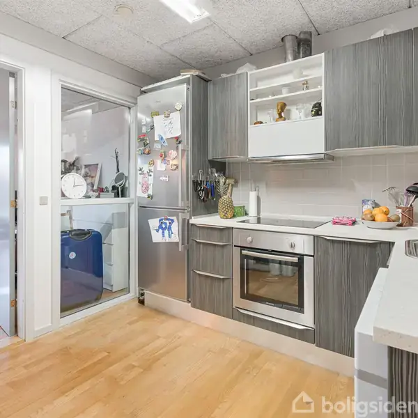 Et moderne køkken med grå skabe og indbygget ovn. Der er en køleskabsmagnet og dekorationer. Ved siden af er der en åben dør til en stue med trægulv og planter.