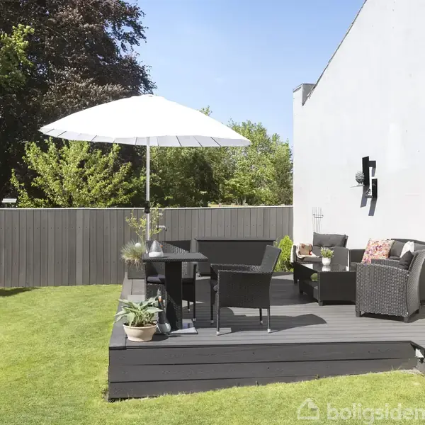 En terrasse med sorte havemøbler og en hvid parasol står ved en hvid mur. Græsplæne og grønne buske omgiver området, der har en hyggelig og solrig atmosfære.