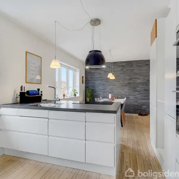 En hvid, moderne køkkenø med mørk bordplade står midt i et lyst rum med trægulv. En stor sort lampe hænger over køkkenøen. I baggrunden ses et spisebord.
