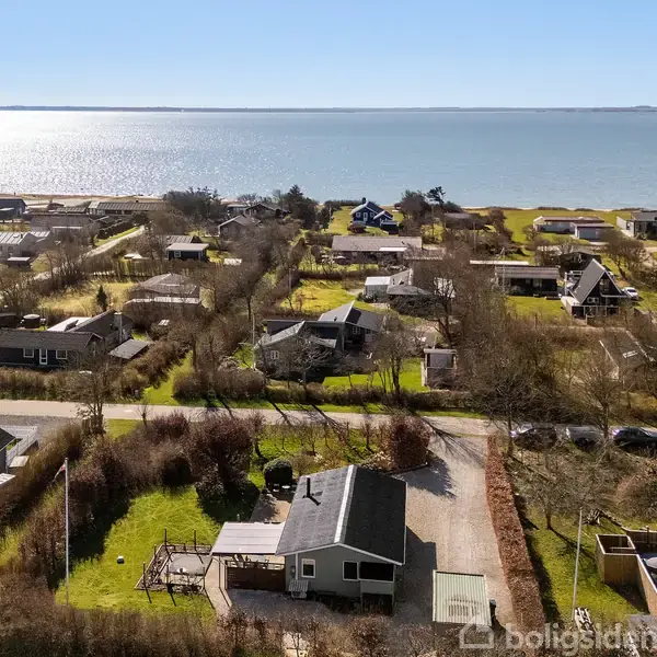 Hus omringet af andre bygninger ligger tæt på en kystlinje. Der er grusveje, grønne græsarealer og vandet ses i baggrunden under en klar blå himmel.