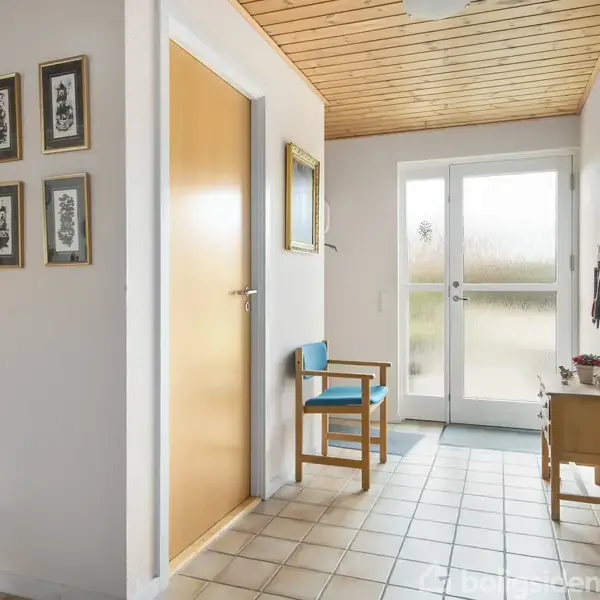 En lys entré med flisegulv. En blå stol står ved siden af en trædør. Væggene har billeder og malerier, mens et lille træbord står ved glasdøren, hvilket leder udenfor.