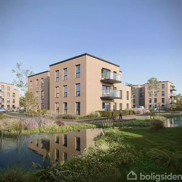 Et moderne lejlighedskompleks spejler sig i en sø omgivet af planter og græs i et boligområde under en klar blå himmel.