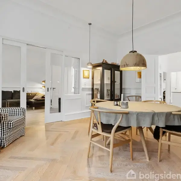 En spisebord med stole står i et lyst rum med parketgulv. Der er en sort- og hvidternet lænestol ved siden af et rundt bord. Store glasdøre fører til et tilstødende rum.