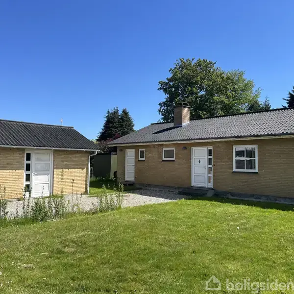 En murstensvilla står stille på en solrig græsplæne forbundet med en tilbygning. Himlen er klar blå, og der er træer i baggrunden.