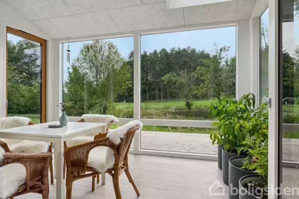 En lys vinterhave med et hvidt bord og flettede stole med pelsbetræk. Store vinduer viser en have med træer. Planter i potter står langs vinduerne.