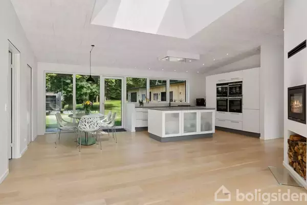 Køkken med kogeø og spisebord. Store vinduer afslører en grøn have udenfor. Hvide skabe, indbygget ovn og pejs er også synlige i det moderne rum.