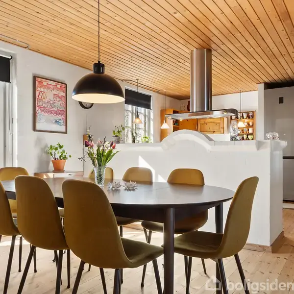 Et moderne spisebord er omgivet af seks stole i et køkken-alrum. En stor lampe hænger over bordet. Køkkenet har åbne hylder, et hvidt halv-væg design og lyst træloft.