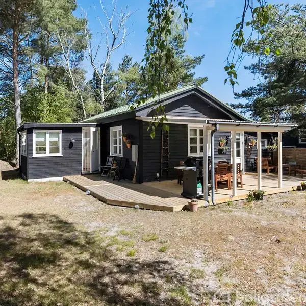 Sort træhus står stille på en lys, åben grund omgivet af træer. Foran huset er der en træterrasse med havemøbler under en overdækket pergola.
