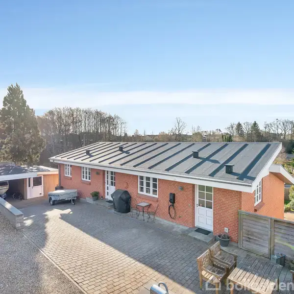 Et enfamiliehus i røde mursten ligger på en solrig dag omgivet af træer og en grusbelagt indkørsel. Foran huset ses en terrasse med havemøbler og grill.