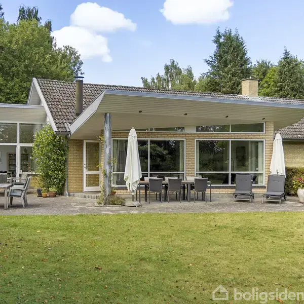 En moderne villa med store vinduer og udendørs terrasse med havestole og parasoller. Den ligger i en grøn have omgivet af træer.