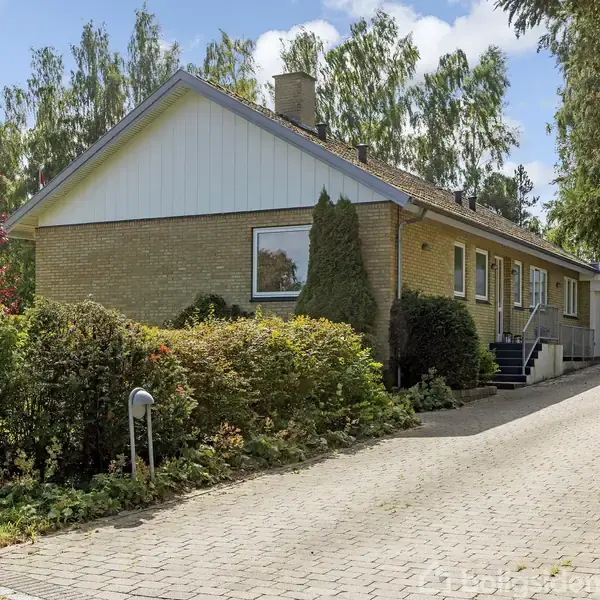 En gul murstensvilla med skorstenspibe, omkranset af buske, træer og velholdt have. Beliggende for enden af en flisebelagt indkørsel, med tilhørende garage i baggrunden.