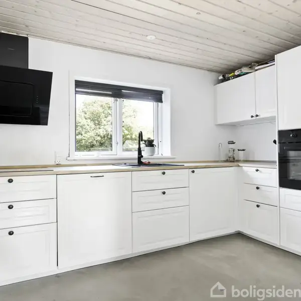 Hvidt køkken med indbyggede skabe og skuffer. Vask og sort emhætte på væggen over en kogeplade. Naturudsigt gennem stort vindue. Moderne, minimalistisk design.