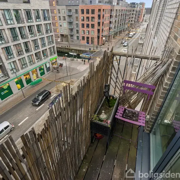 Lille altan med træhegn og to lilla stole, set ovenfra. Befinder sig på en etagebygning med udsigt over en byvej omgivet af lejlighedskomplekser.