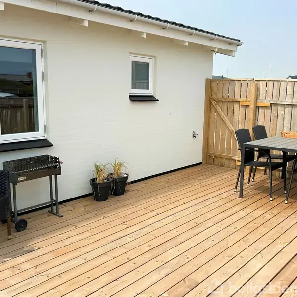 Terrasse med trægulv, bord og stole ved siden af en grill. Planter står ved en hvid husvæg med vinduer, omkranset af et hegn, i et lyst udeområde.