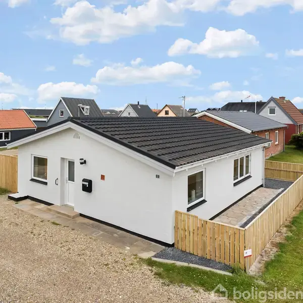 Et hvidt étplanshus med sort tag står stille på en grusvej. Huset er omgivet af et lavt træhegn, og der er flere lignende huse i baggrunden under en blå himmel med skyer.