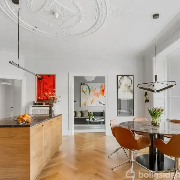 Køkken med træbordplade og køkkenø fyldt med appelsiner. Rundt spisebord med brune stole, omgivet af moderne kunst på væggene, i et rum med stukloft og store vinduer.