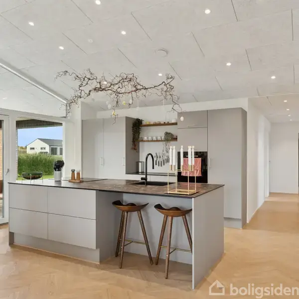 Et moderne, lavt hus i lysebrune mursten står på en flad grund med grusbelægning. Huset har flere vinduer og en sorte tagsten. Området er roligt og boligagtigt.