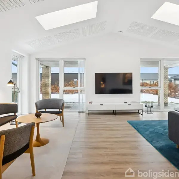 Stue med grå sofaer og bord, placeret på trægulv; en fladskærm hænger på hvid væg. To store vinduer i baggrunden giver udsigt til en terrasse.