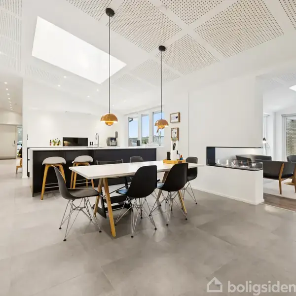 Spisebord i moderne, lyst køkken-alrum. Seks stole omgiver bordet under to hængende lamper. I baggrunden en pejs og stue med sofaer og store vinduer.