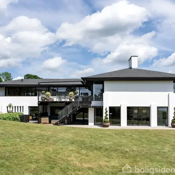 En moderne, hvid villa med store glaspartier står på en velplejet græsplæne. Der er en terrasse med trapper, og landskabet omkring er grønt med få træer og en blå himmel.