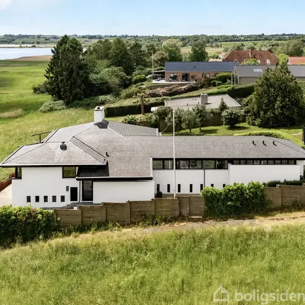 Et moderne, hvidt hus med fladt tag står i en frodig, grøn landskab. Huset har store vinduer, og der er en trampolin i haven. I baggrunden ses en sø og flere træer.