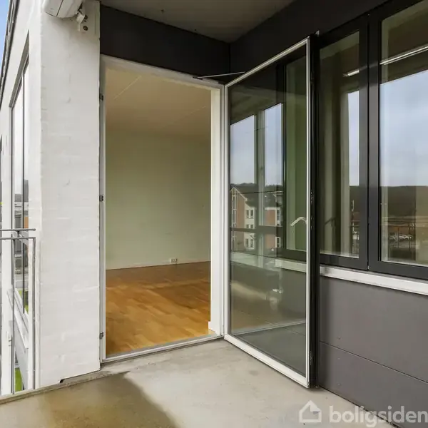 Altan forbinder til lejlighed via skydedør; trægulv indendørs. Moderne bygning med hvid murstensfacade og glasparti. Udsigt over kvarter, græsplæner og nabobygninger under en lys himmel.