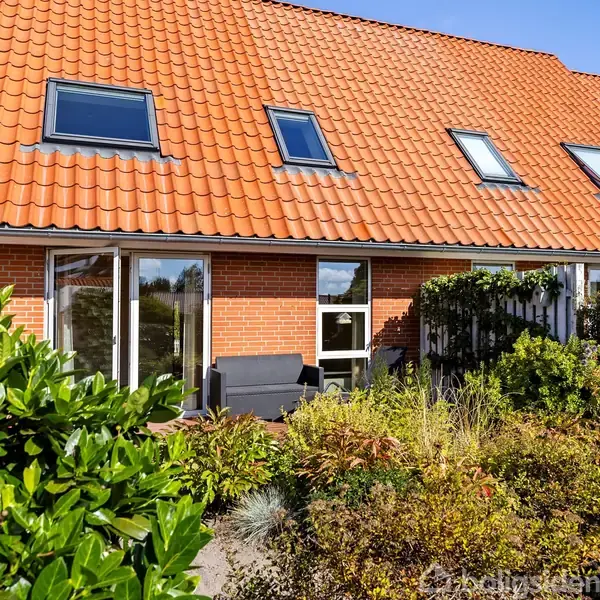 Et rækkehus med rødt tegltag og flere ovenlysvinduer står i sollys. Foran huset er en terrasse omkranset af frodig vegetation og buske.