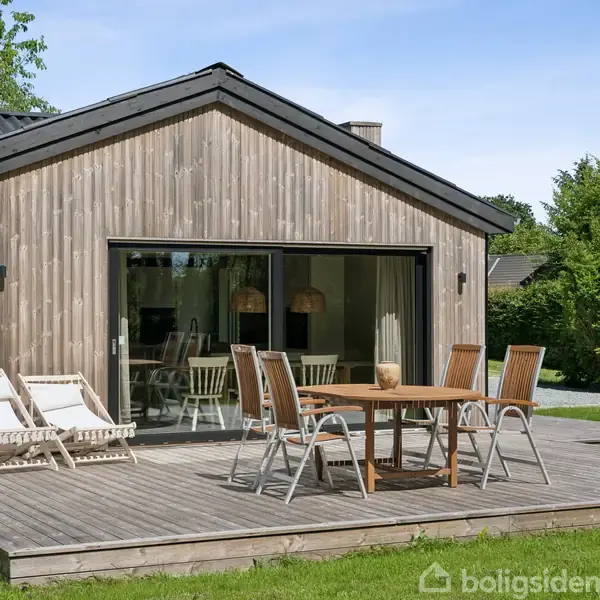 Et træhus med store glasskydedøre. Foran står et rundt træbord med stole og solsenge. Omgivet af terrasse og grøn vegetation i baggrunden.