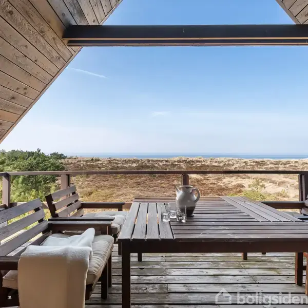 Bord med bænk og stole står på en overdækket træterrasse. Bordet har en vase, og terrassen har udsigt over et landskab med træer og hav i baggrunden.
