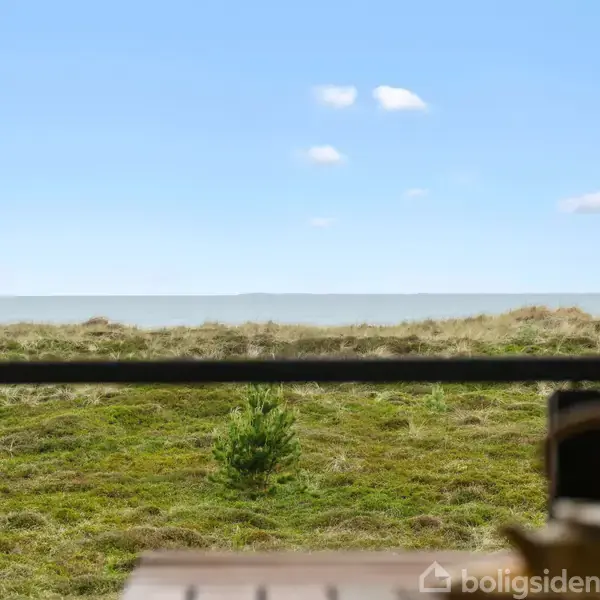 Bord står stille foran en vidstrakt græsbevokset klitlandskab. Over horisonten ses hav og blå himmel med spredte skyer. Ankeret i forgrunden skaber forbindelse mellem natur og indretning.