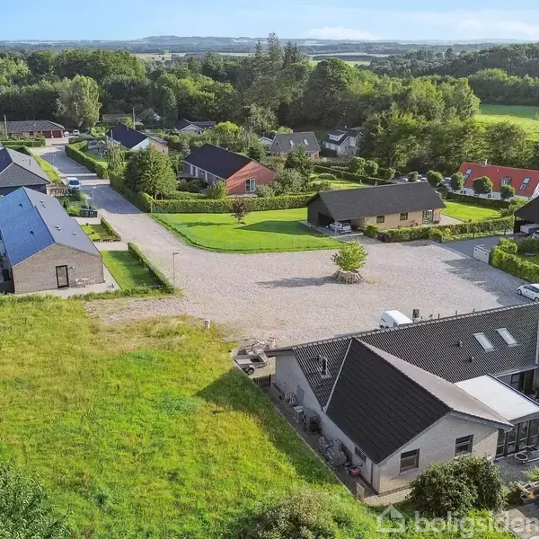 Flere moderne enfamiliehuse med sorte tage står langs en stille vej i en grøn, landlig landsbyomgivelse. Træer omgiver området, og velplejede haver kan ses ved husene.