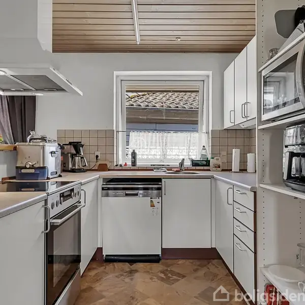 Køkken med hvide skabe og en sort komfur, kaffemaskine og mikrobølgeovn. En opvaskemaskine er under et vindue med flisebelagt backsplash. Træloft og beige flisegulv i hjemligt interiør.