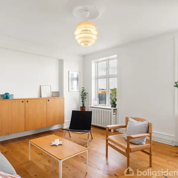 Stue indrettet med sofa, lænestole og sofabord. Vægskab ophængt, planter dekorerer rummet. Trægulv og hvidmalede vægge skaber lys atmosfære, store vinduer giver naturligt lys.