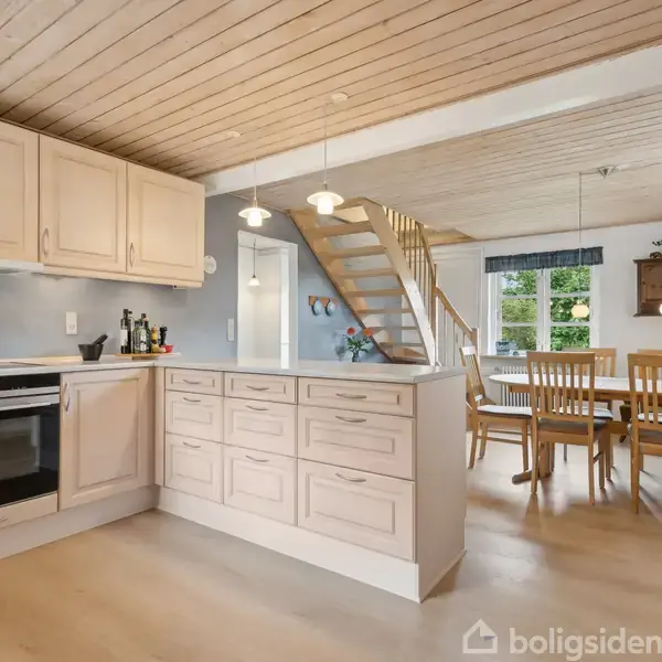 Et lyst køkken med træskuffer og skabe, der står ved siden af en åben trappe. Et spisebord med træstole ses i baggrunden nær et stort vindue.