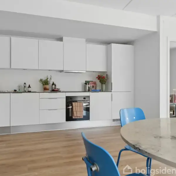 Et moderne køkken med hvide skabe, hvor en ovn og kogeplade er integreret. Der står planter og køkkenredskaber på bordpladen. Et rundt marmorbord og blå stole står foran.